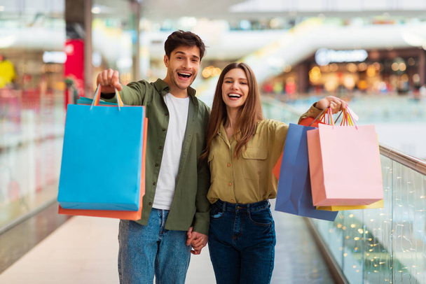 Gli sposi allegri che mostrano borse della spesa alla macchina fotografica in piedi nel centro commerciale - Foto, immagini