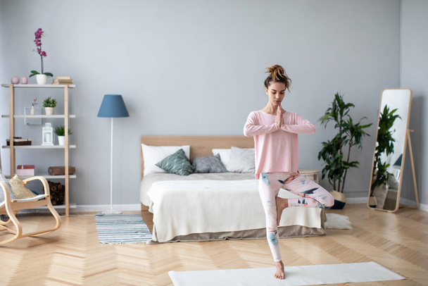 Young beautiful woman with hair bun in sportswear practicing yoga at home, doing Vrksasana exercise on one leg, hands in prayer namaste gesture, Tree pose. - Foto, afbeelding