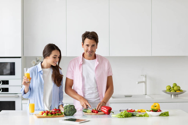 Healthy meal concept. Handsome man cooking fresh vegetable salad, girl standing with a glass of orange juice in the kitchen. - Zdjęcie, obraz