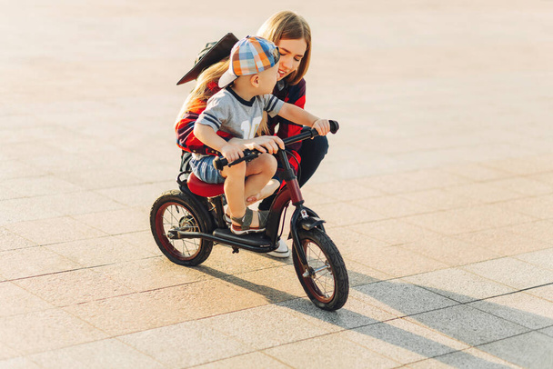 szczęśliwe dziecko jeździ na rowerze, podczas gdy jego matka idzie obok niego. Szczęśliwa matka uczy syna jeździć na rowerze w parku w słoneczny dzień. Pojęcie rodziny, macierzyństwa i rekreacji - Zdjęcie, obraz
