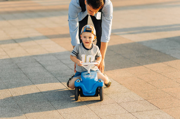 Dad teaches his little son to ride a children's car in the park, keep balance, have fun with the family. Father and son in the park, the boy is driving a children's car. Father's day - Zdjęcie, obraz