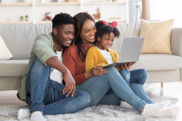 Χαρούμενη αφροαμερικάνικη οικογένεια τριών ατόμων που χαλαρώνουν με φορητό υπολογιστή στο σπίτι - Φωτογραφία, εικόνα