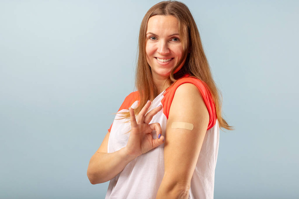 Successful Covid-19 vaccination. Vaccinated woman is gesturing okay and showing arm with adhesive bandage after vaccine injection. COVID-19 vaccination concept. - Фото, зображення