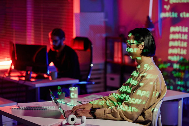 Σύγχρονη νεαρός μηχανικός λογισμικού κάθεται στο χώρο εργασίας μπροστά από φορητό υπολογιστή - Φωτογραφία, εικόνα