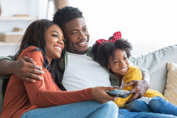 Домашнее времяпровождение. Счастливая афроамериканская семья из трех человек смотрит телевизор дома - Фото, изображение