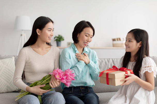 Щаслива азіатська дівчинка, яка дарує своїй матері й бабусі загорнуті подарунки й квіти, вітаючи їх з відпусткою вдома. - Фото, зображення