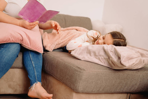 Przycięte zdjęcie matki siedzącej na kanapie i trzymającej wieczorem książkę czytającą bajkę dla śpiącej córki., - Zdjęcie, obraz