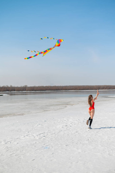 Caucasian woman in a red swimsuit launches a kite on the seashore in winter.  - 写真・画像