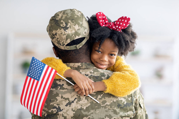 Χαριτωμένο μικρό μαύρο κορίτσι κρατώντας αμερικανική σημαία και αγκαλιάζει τον μπαμπά στρατιώτη - Φωτογραφία, εικόνα