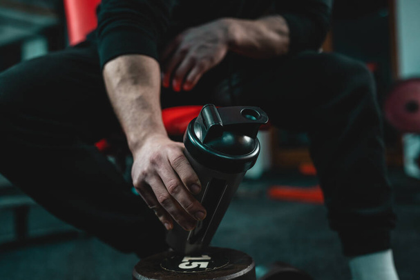 Close-up na mão de homem caucasiano desconhecido segurando shaker suplemento escuro enquanto sentado no ginásio durante o treinamento espaço de cópia foco seletivo - Foto, Imagem