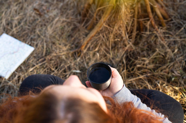 Foto vista superior de una mujer al aire libre sentada en la hierba seca con las piernas cruzadas sosteniendo una taza de té caliente cerca de la hoguera. - Foto, Imagen
