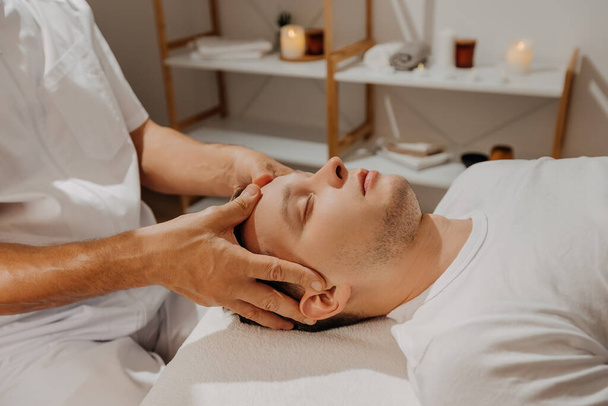 Hombre joven relajante durante el masaje de cabeza profesional en el salón SPA. Tratamiento manual de migraña. Concepto de bienestar, cuerpo y salud - Foto, imagen