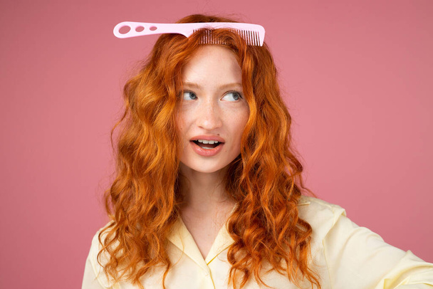 Picture of lovely redhead girl with fine hair comb on her head posing for the camera over pink background, cut hair concept. - Photo, image