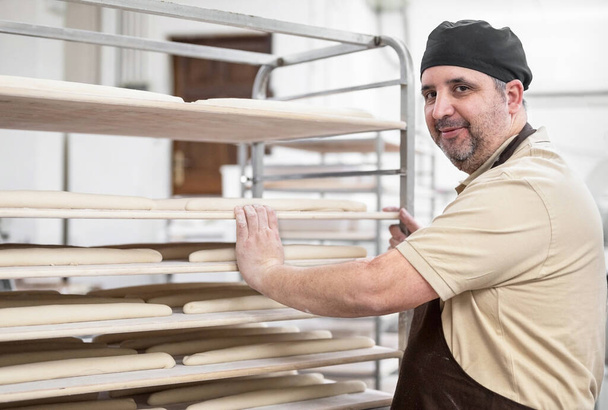 Confident baker posing with rack of fresh bread loaf at bakery. - Photo, Image