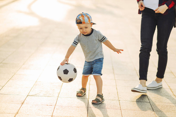 Äiti ja poika pelaavat jalkapalloa yhdessä ulkona puistossa, pieni poika potkaisee jalkapalloa, lapsi oppii pelaamaan jalkapalloa. Perhe pelaa jalkapalloa yhdessä puistossa - Valokuva, kuva