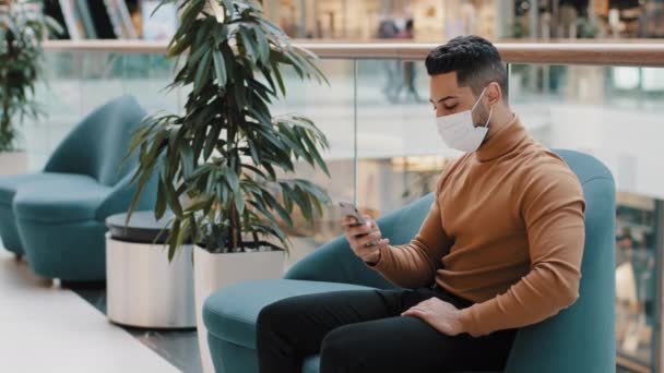 Young man in medical mask sits indoors typing message on social network on smartphone sends email via phone using mobile application for ordering in internet store web surfing on online dating site - Footage, Video