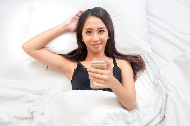 Азиатка, лежащая на кровати, используя мобильный телефон - Фото, изображение