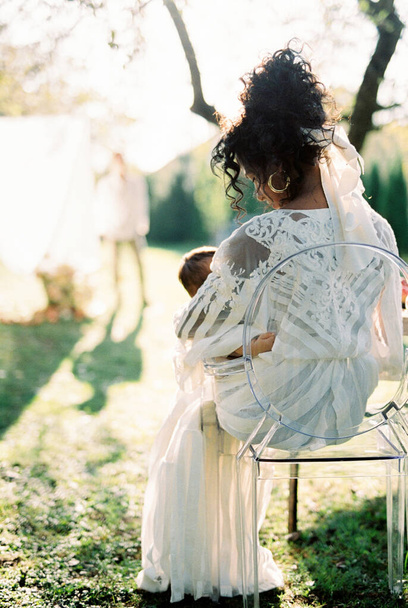 Donna con un bambino tra le braccia si siede su una sedia in un giardino verde - Foto, immagini