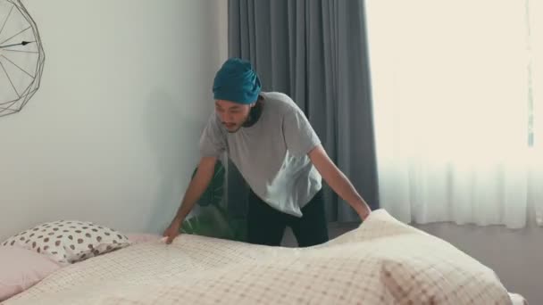 Een aantrekkelijke Aziatische man regelt kussens in de slaapkamer voor het uitgaan. De ochtendzon schijnt door het raam thuis. - Video