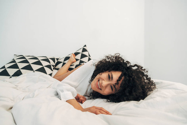Χαμογελώντας παιχνιδιάρα γυναίκα μικτής φυλής ξαπλωμένη στο κρεβάτι κοιτάζοντας την κάμερα. Αντιγραφή χώρου. - Φωτογραφία, εικόνα