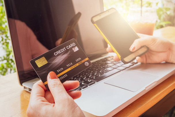 Κοντινό πλάνο κάποιου κρατώντας με το χέρι πιστωτική κάρτα κατά τη διάρκεια της χρήσης laptop και κινητό τηλέφωνο για online αγορές. Online αγορές είναι η διαδικασία αγοράς αγαθών και υπηρεσιών από εμπόρους μέσω του Διαδικτύου. - Φωτογραφία, εικόνα