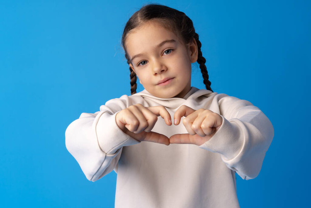 Счастливая маленькая девочка делает форму сердца руками на синем фоне. - Фото, изображение