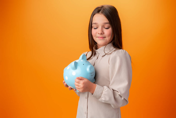 Teen girl with piggy bank on orange color background. - Фото, изображение