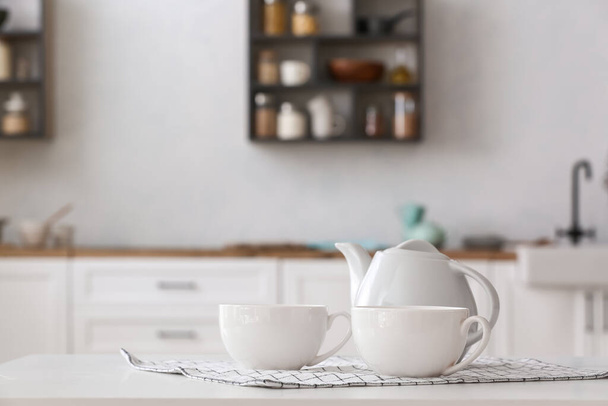 Чайник с чашками и салфеткой на столе в светлой кухне - Фото, изображение