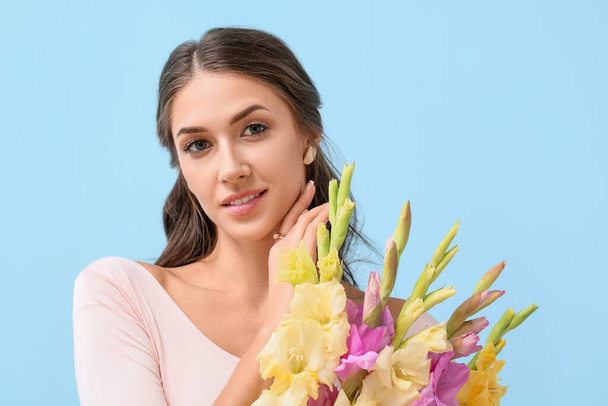 Mujer joven con la mano cerca de la cara y flores de gladiolo sobre fondo de color - Foto, Imagen