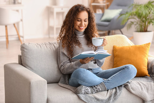 Νεαρή Αφρο-Αμερικανίδα γυναίκα με φλιτζάνι τσάι ανάγνωση βιβλίο στον καναπέ στο σπίτι - Φωτογραφία, εικόνα