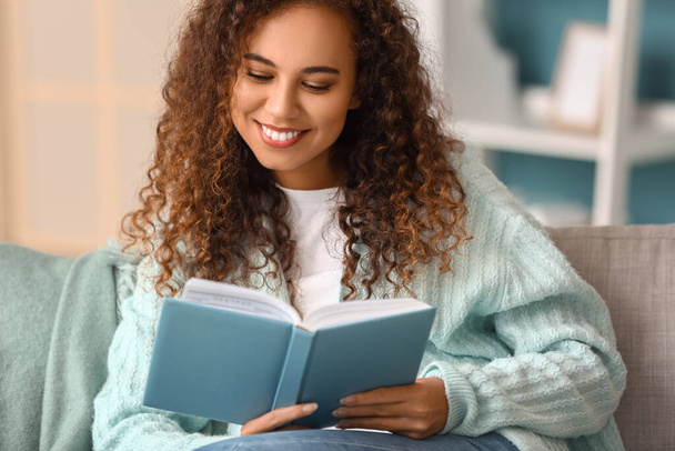 Jonge Afro-Amerikaanse vrouw in warm vest leesboek op de bank thuis - Foto, afbeelding
