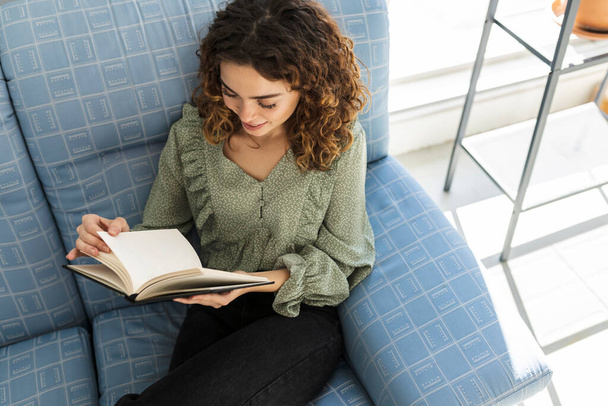 nízký úhel pohledu na kudrnaté vlasy žena čtení knihy na pohovce, hezká běloška, baví doma - Fotografie, Obrázek