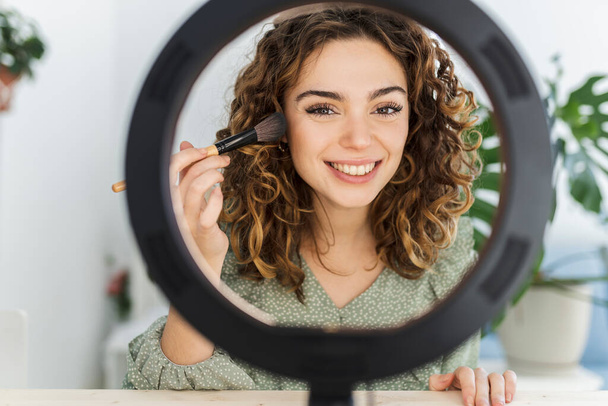 krullende vrouw zet op make-up in spiegel met licht thuis - Foto, afbeelding