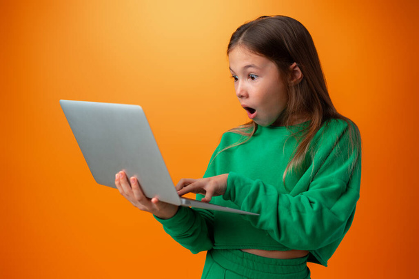 Portrait of a cheerful young teen girl holding laptop computer against orange background - Foto, Imagem