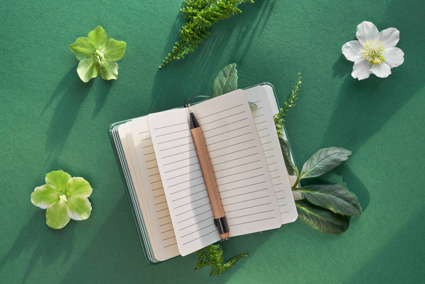 Notebook in with exotic green plants and flowers. Fern leaves, green and white Helleborus flowers. Creative natural flat lay, top view on dark green paper background, long shadows. - Fotó, kép