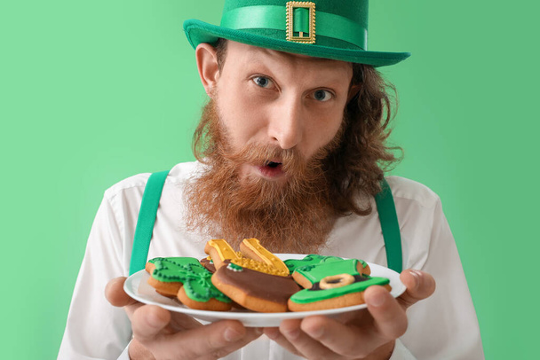 Hombre barbudo sorprendido sosteniendo placa con galletas sobre fondo verde, primer plano. Celebración del Día de San Patricio - Foto, Imagen