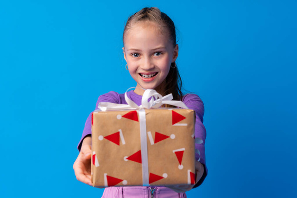 Happy little girl with long hair holding a gift box on a blue background - Zdjęcie, obraz