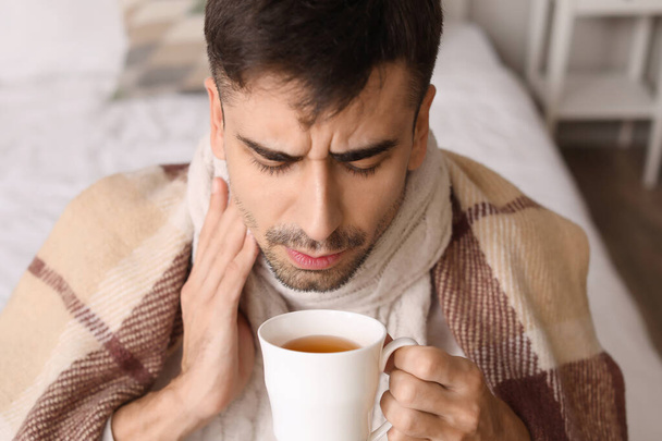 Kranker junger Mann mit Halsschmerzen trinkt zu Hause heißen Tee - Foto, Bild
