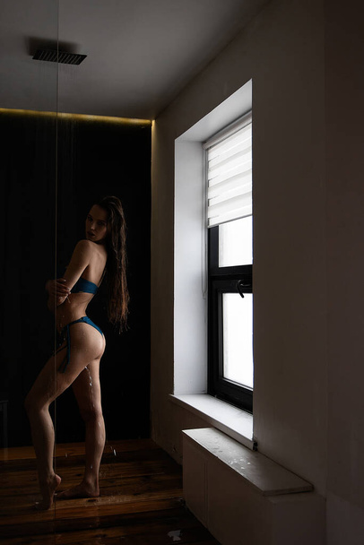 Sexy szczupła brunetka z długimi mokrymi włosami w niebieskim stroju kąpielowym z gołymi ramionami i brzuchem dotykającym ciała patrząc w dół na czarnym tle - Zdjęcie, obraz