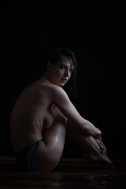 Вид сбоку полное тело молодой стройной сексуальной барышни с темными волосами в трусиках-бикини без бюстгальтера, сидящей на деревянном полу в душе и покрывающей обнаженную грудь - Фото, изображение