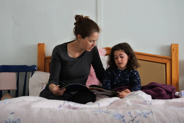 Dorosła matka (kobieta w wieku 30-40 lat) czytająca opowiadanie z książki córce (kobieta w wieku 08 lat) w łóżku. - Zdjęcie, obraz