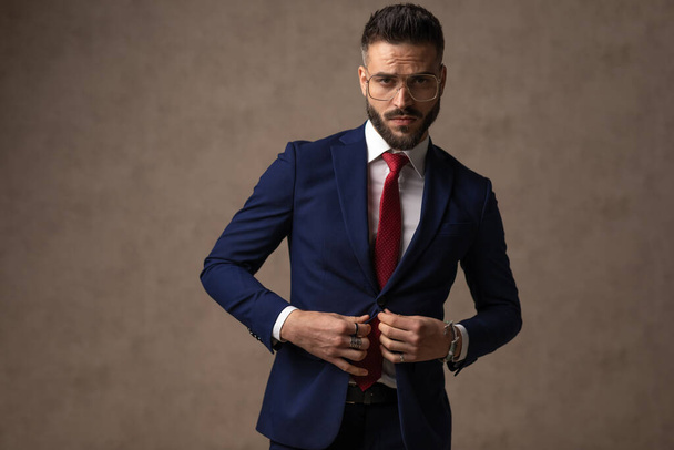 junger sexy Geschäftsmann arrangiert seinen Anzug, trägt eine Brille und macht ernst gegen Tapete - Foto, Bild