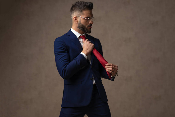 boční pohled na pohledného podnikatele, jak si upravuje kravatu a tváří se jako macho proti tapetě - Fotografie, Obrázek