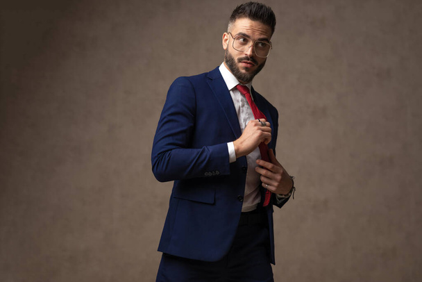 sexy hombre de negocios arreglando su corbata, mirando hacia otro lado, usando anteojos y posando contra el papel pintado - Foto, imagen