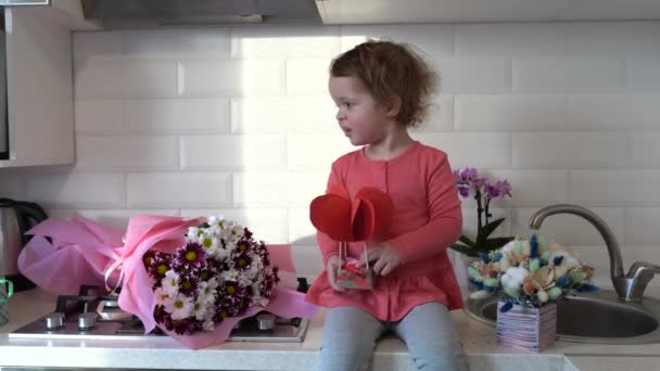 Szczęśliwych Walentynek Słodkie dziecko dziewczyna gra z wystrojem i siedzi na stole w kuchni z kwiatami w domu - Materiał filmowy, wideo