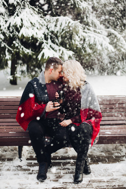 Bankta oturan ve orman kar yağışı gülümseyen aşık çift. - Fotoğraf, Görsel