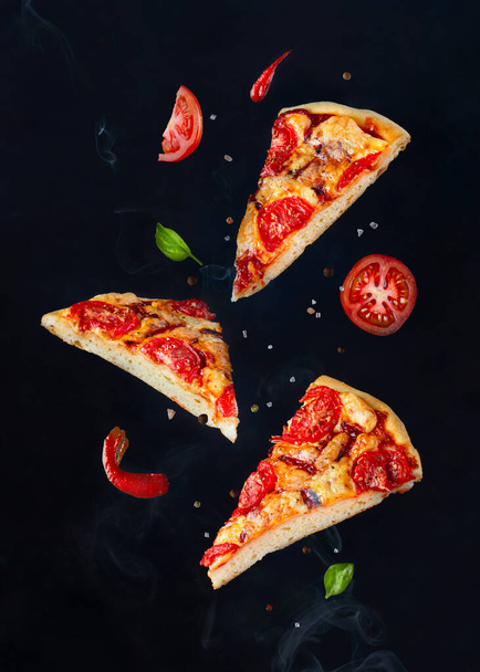 Pieces of pizza flying with herbs, sauce and spices on a dark background with smoke - Photo, image