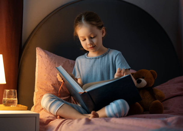 Cute little girl is reading a book in her bedroom. - Foto, imagen