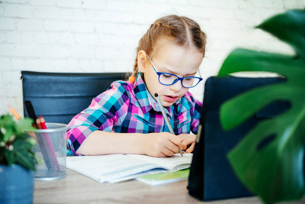 A szemüveges kislány jegyzetel a jegyzetfüzetébe, miközben otthon tanul az interneten. - Fotó, kép