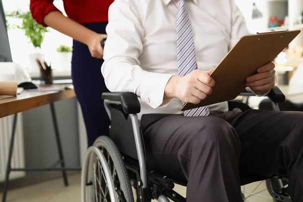 Man zit in rolstoel met klembord, vrouw rijden baas in kantoor - Foto, afbeelding